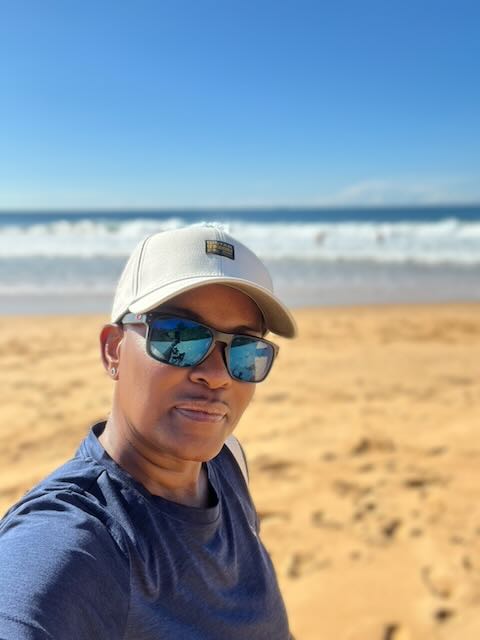 Samantha Haynes taking a beach selfie wearing a cream cap and blue mirrored sunglasses, with golden sand, rolling waves and a clear blue sky in the background