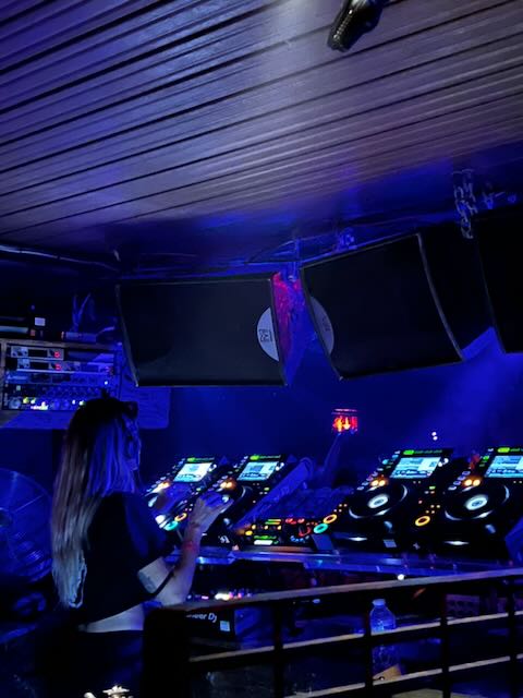 A female DJ performing at a club, working a DJ setup with multiple decks, bathed in blue UV lighting with suspended speakers and screens above.