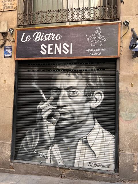 A striking monochrome mural of a cigarette-smoking figure covers the shuttered entrance of Le Bistro Sensi, a tapas and small plates restaurant, signed by the artist Si. Berlana on a quiet city street