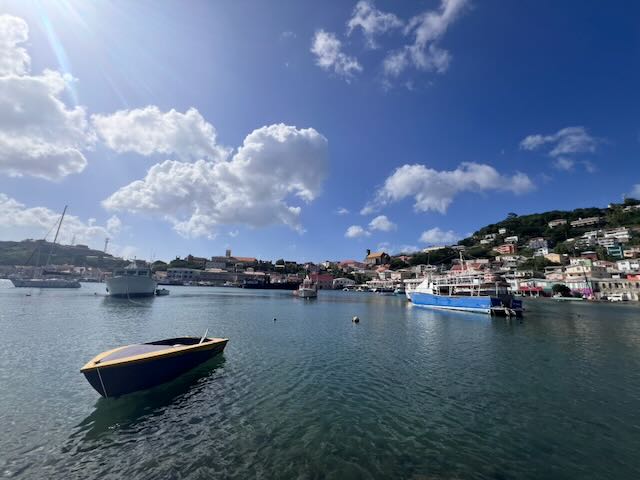 A small wooden boat bobs on the calm turquoise waters of St. George's Harbour, Grenada, with colourful vessels moored nearby and the sun-drenched hillside town climbing into a bright blue sky with scattered clouds.