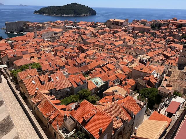 A sea of terracotta rooftops sweeps across Dubrovnik's medieval Old Town, viewed from the city walls, with the deep blue Adriatic and the forested island of Lokrum stretching out beyond.