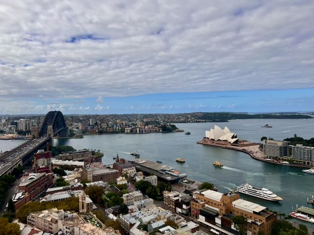 Aerial view of Sydney Harbour, Australia, showing the Sydney Opera House and Sydney Harbour Bridge spanning the water, with the city spreading out beneath a partly cloudy sky.