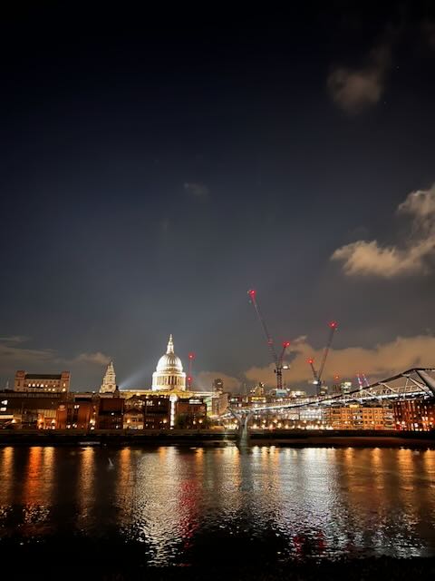 St Paul's Cathedral glowing against the night sky, reflected in the River Thames, with the Millennium Bridge stretching across the water and construction cranes dotting the horizon.